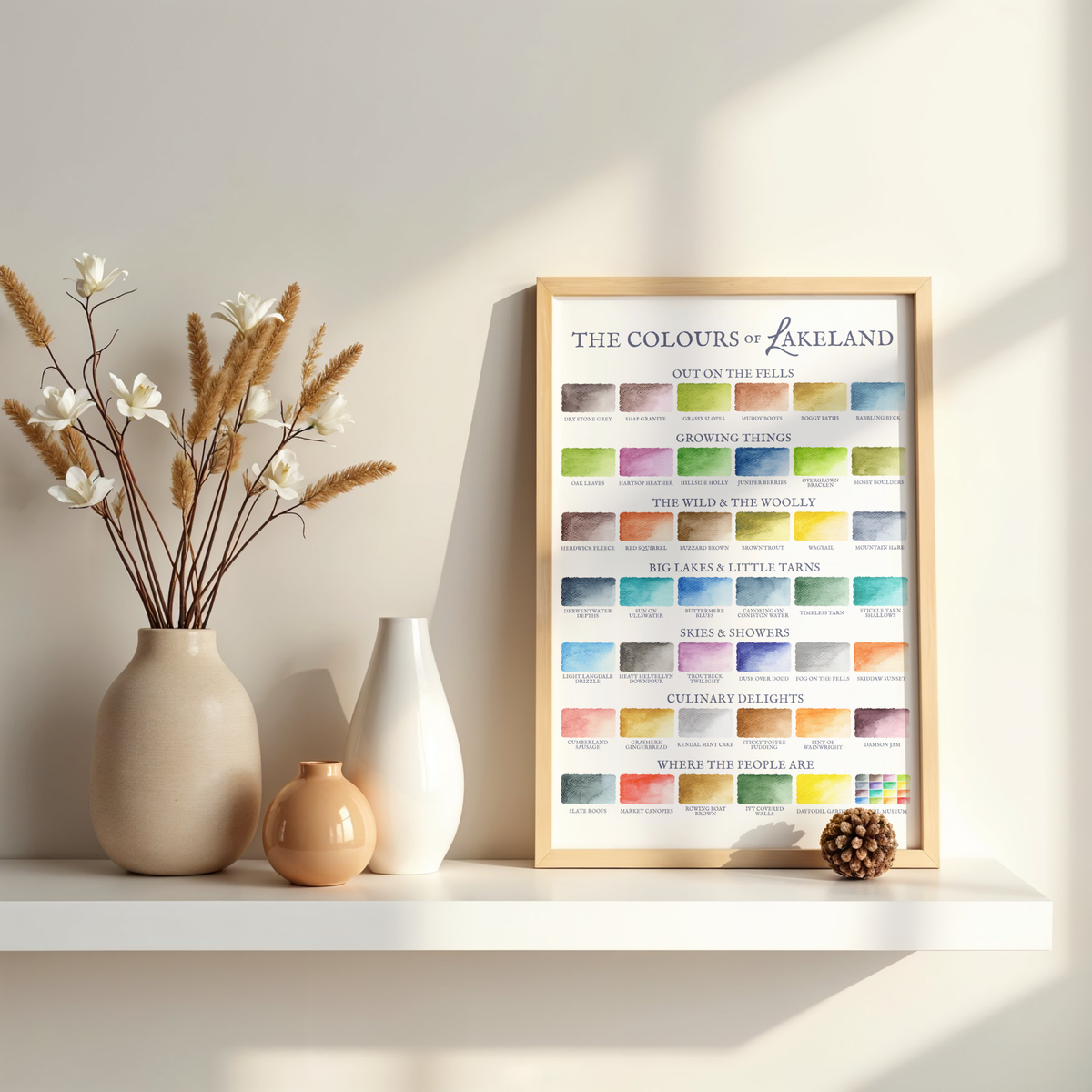 Framed artwork titled 'The Colours of Lakeland' on a shelf with decorative vases and plants.