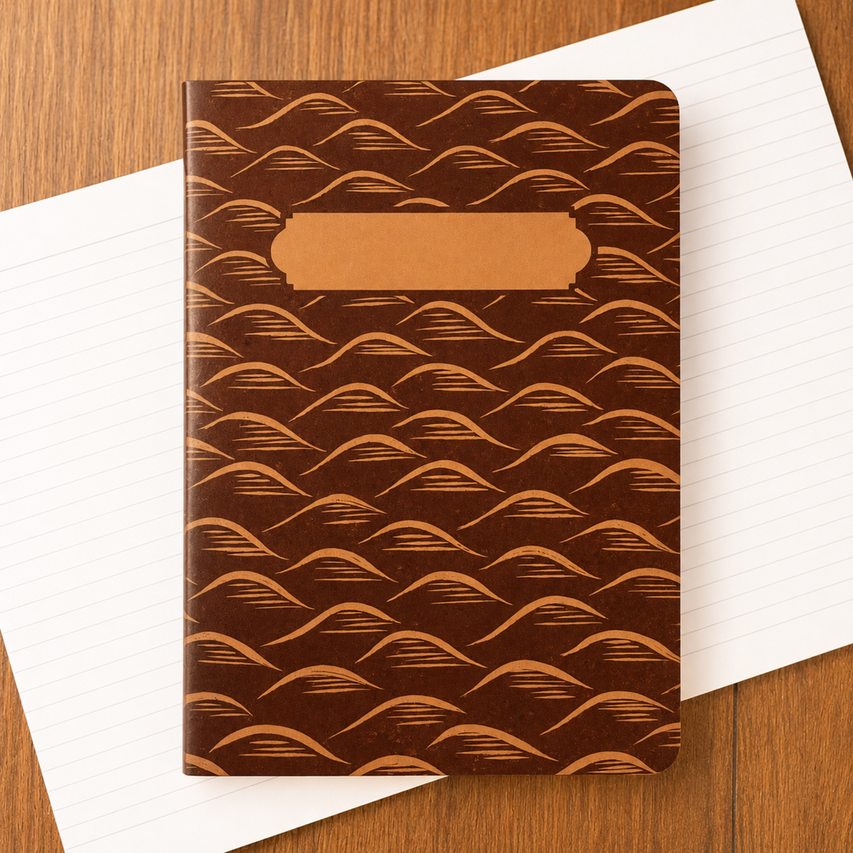 A brown notebook with a repeating tan mountain pattern and a blank tan label area on the front, placed on top of white lined paper over a wooden surface.