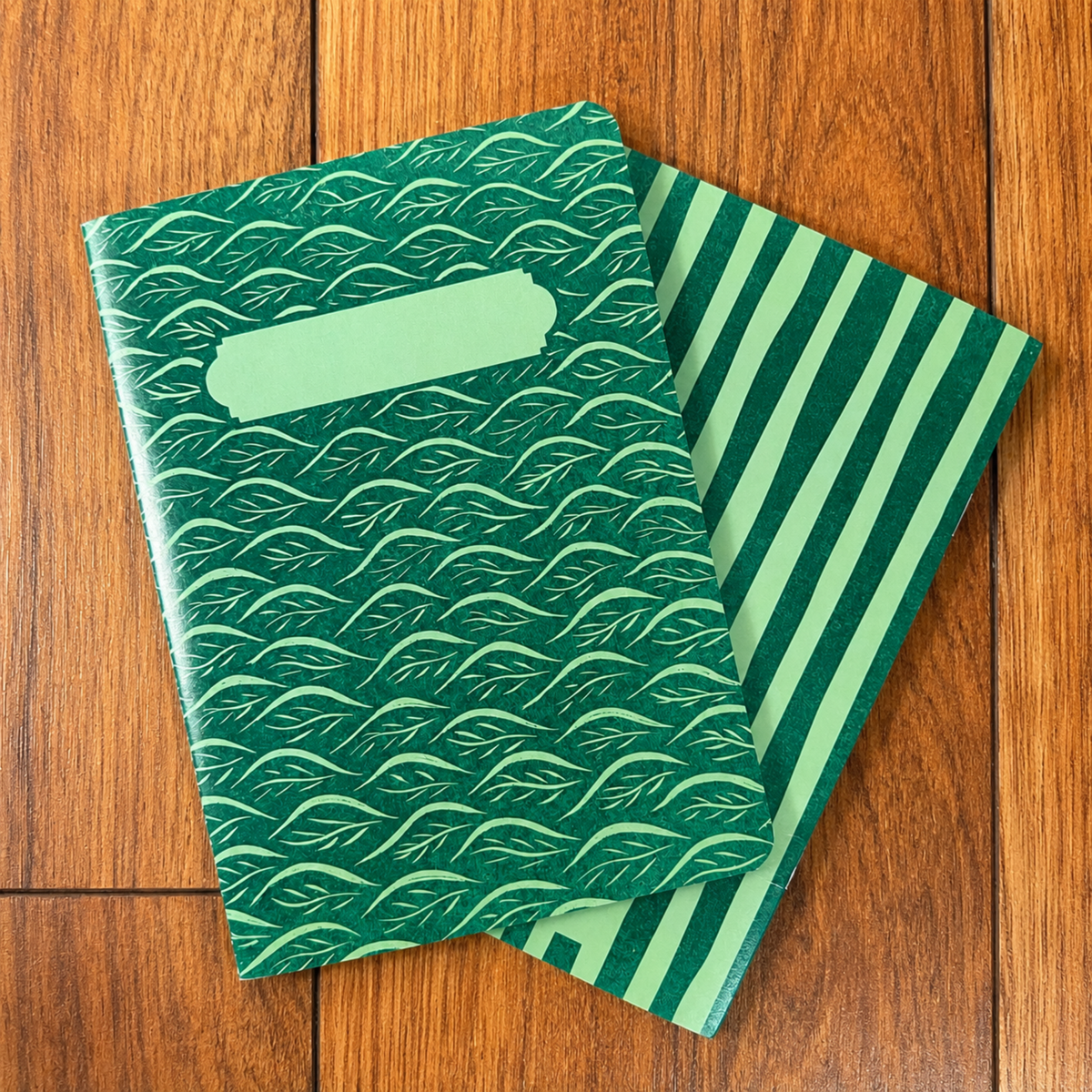 Two green notebooks stacked on a wooden surface. The top notebook has a dark green background with a light green repeating leaf pattern and a blank light green label slot near the top.