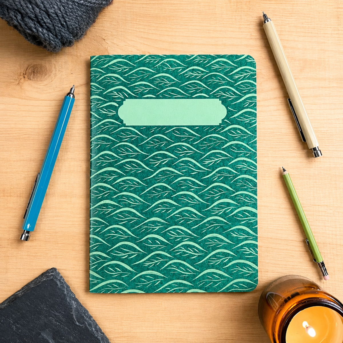 A teal-green notebook with a repeating leaf pattern and a blank label slot near the top, styled on a light wood desk with pens and a candle.