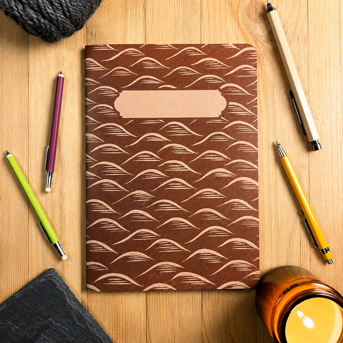A brown Lino Print style notebook with a beige wavy pattern and a blank label on the front, styled on a wooden tabletop with pens, a candle, and a black cloth.