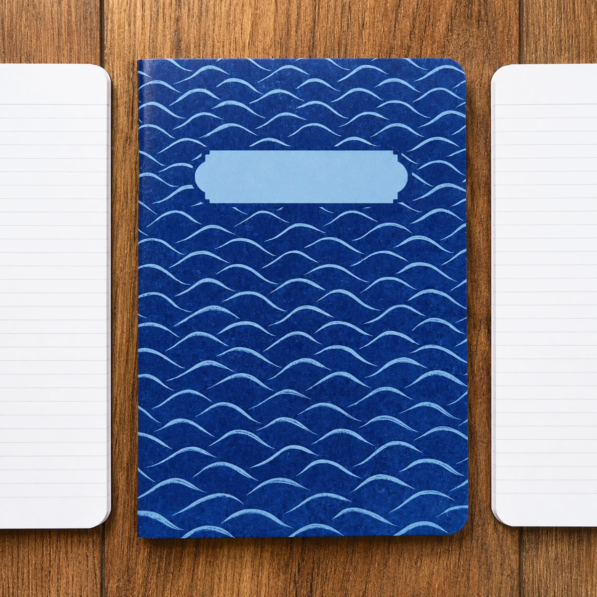 A blue hardcover notebook with a repeating wave pattern and a blank light blue label area on the front, placed on a wooden surface with two white lined notebooks on either side.