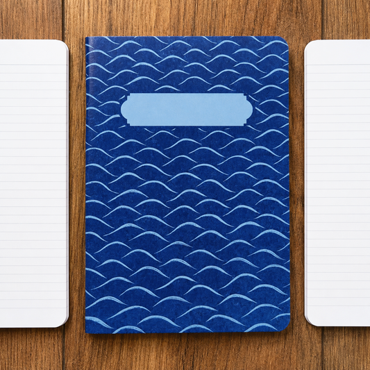 A blue hardcover notebook with a repeating wave pattern and a blank light blue label area on the front, placed on a wooden surface with two white lined notebooks on either side.