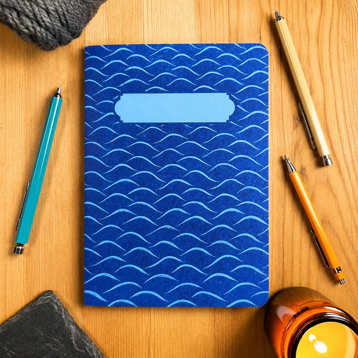 A blue notebook with a lino print style wavy pattern and a blank light blue label on the front, styled on a wooden desk with pens and a candle