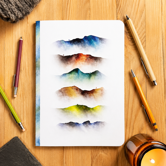 A white notebook with layered watercolour mountain designs in various colours, displayed on a wooden desk with pens, a candle, and a knit scarf.