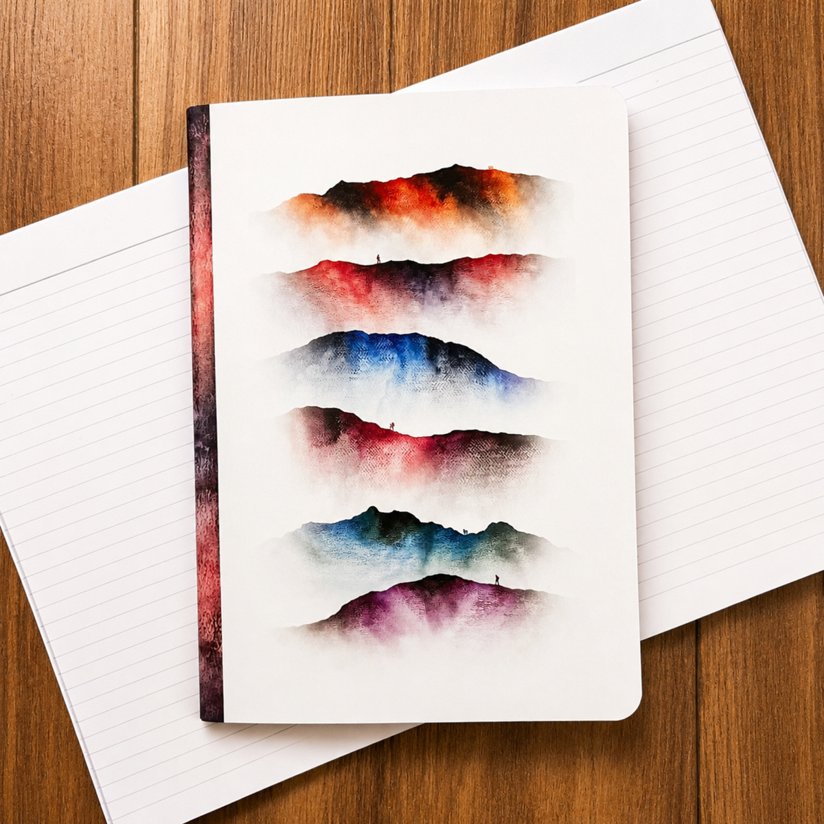 A white notebook with six mountain watercolour designs in red, blue, green, and purple, displayed on top of a lined notebook over a wooden surface.