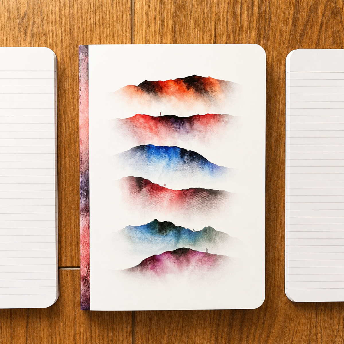 A white notebook with abstract watercolour mountain designs in red, blue, and purple, displayed on a wooden table between two lined loose-leaf pages.