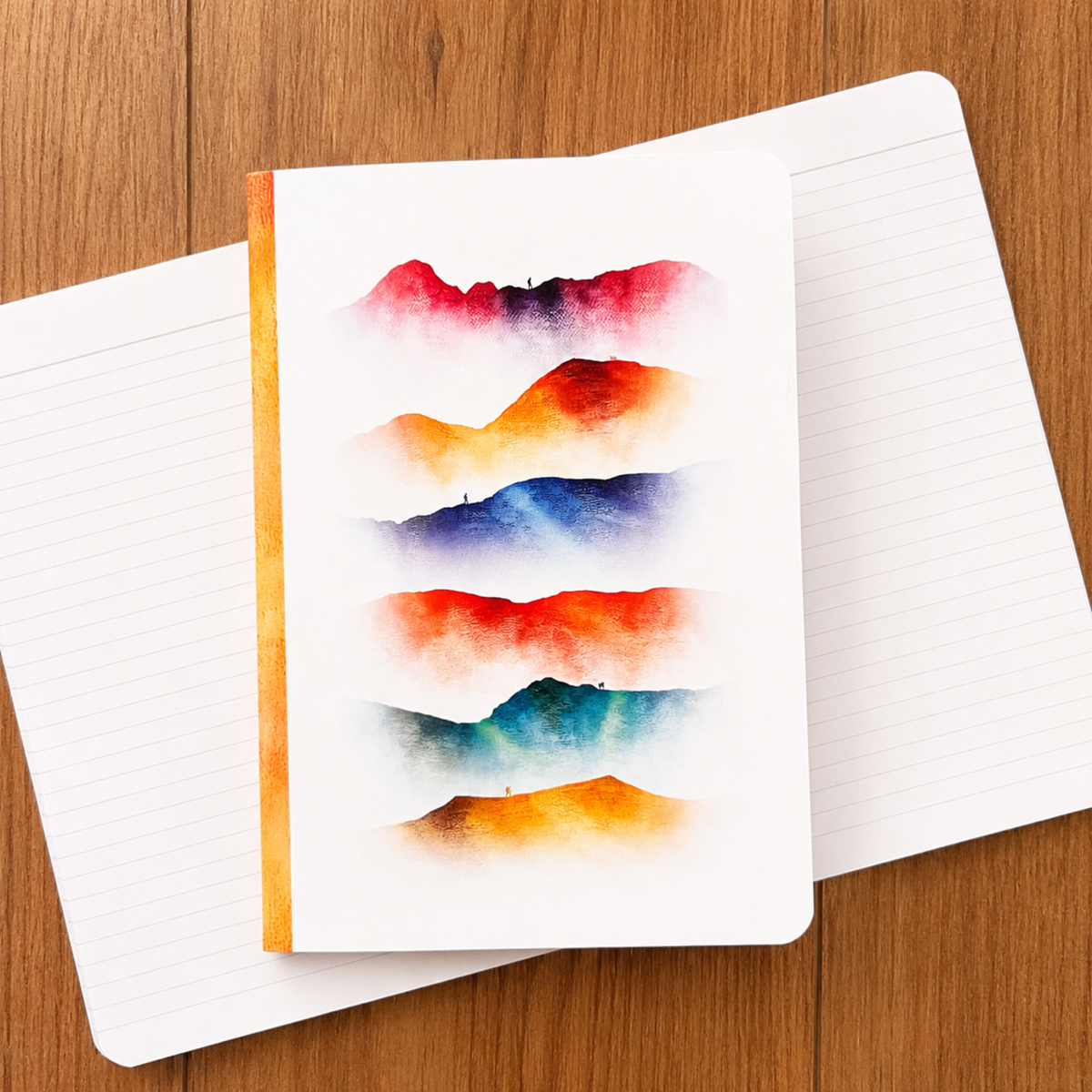 A white lined notebook with an abstract watercolour cover design featuring layered red, orange, blue, and teal mountain-like shapes, placed on top of another open notebook on a wooden surface.