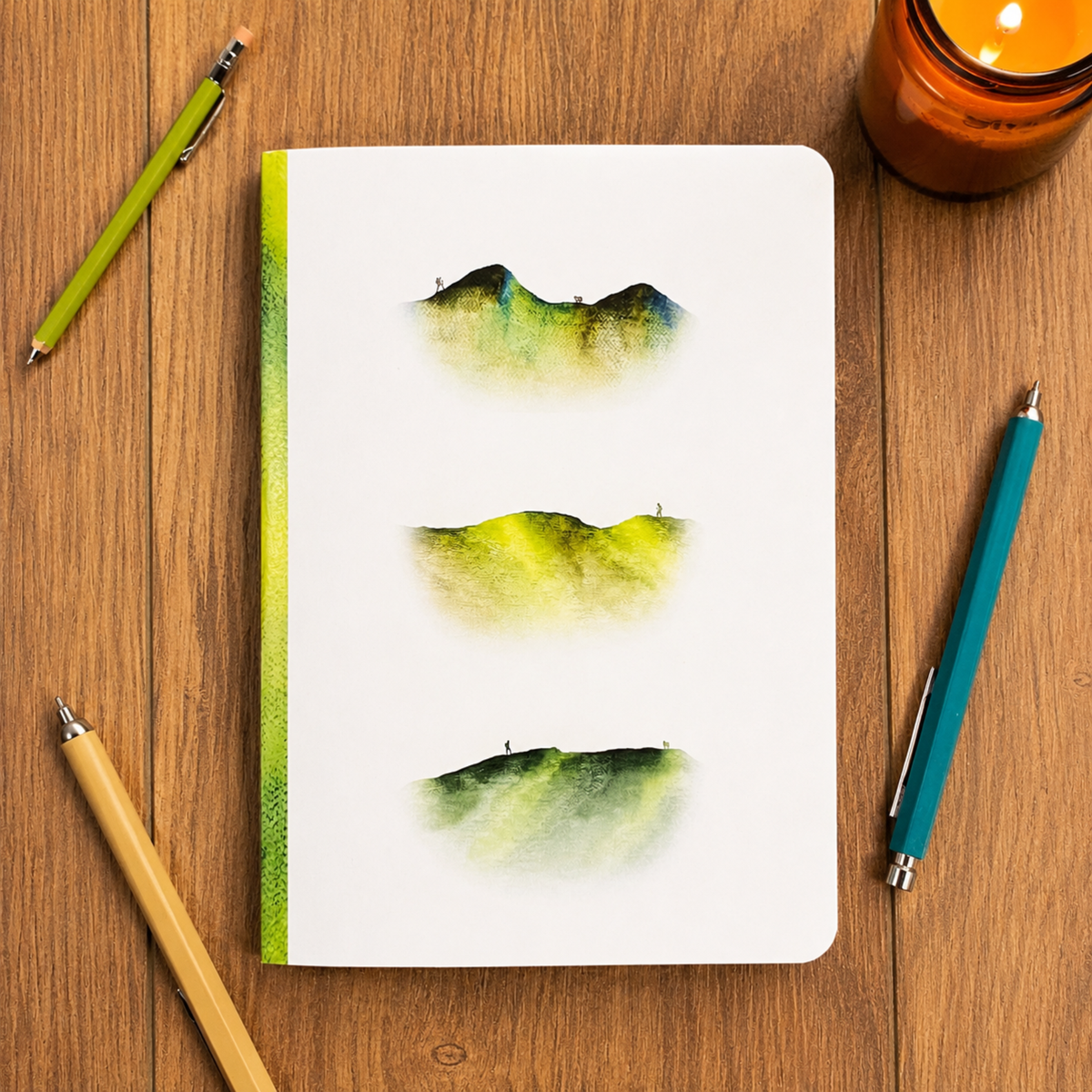 A white notebook with three watercolor mountain landscape prints in green and yellow hues, displayed on a wooden desk with pens and a candle.