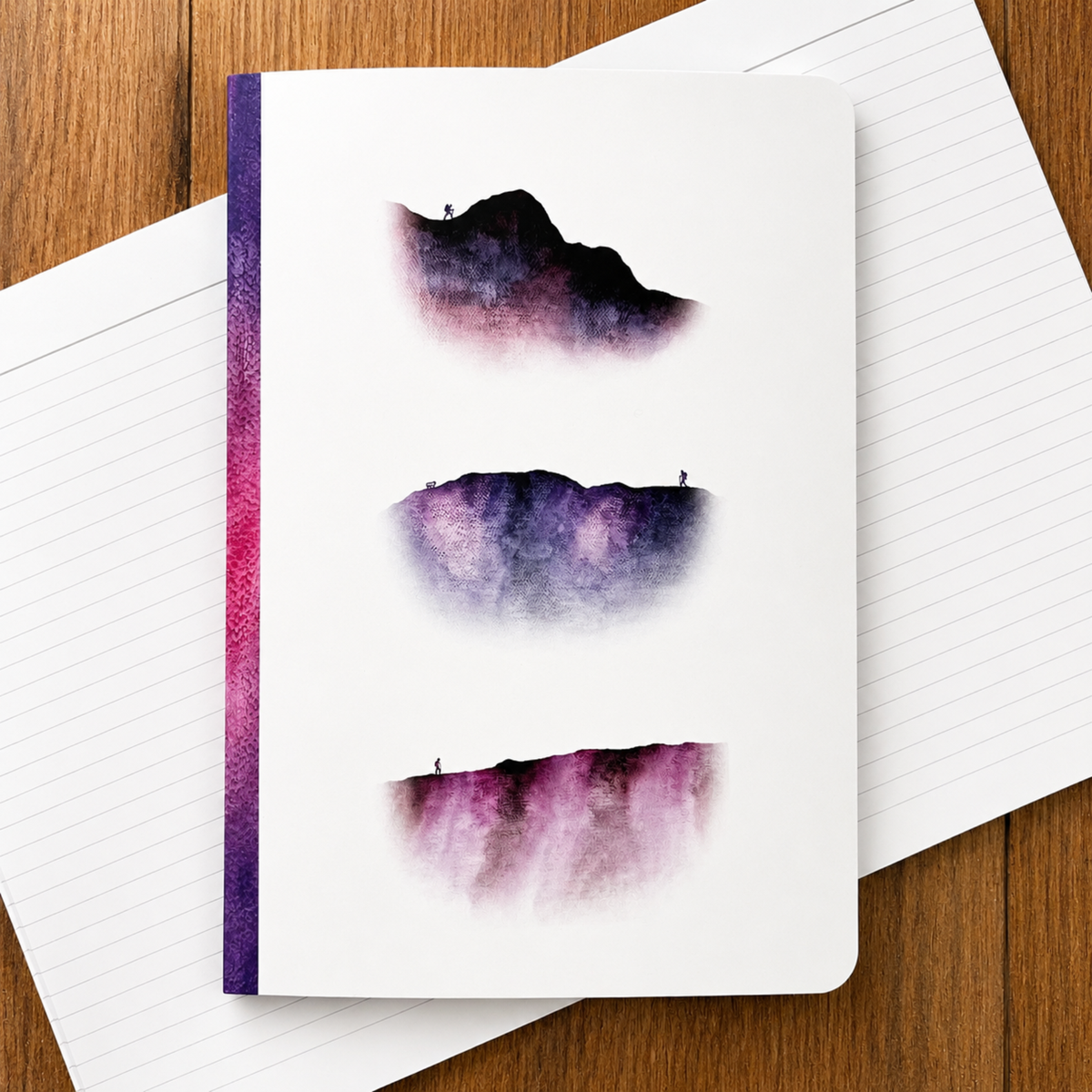 A white notebook with three abstract watercolor mountain designs in purple, pink, and black, displayed on top of open lined paper over a wooden surface.