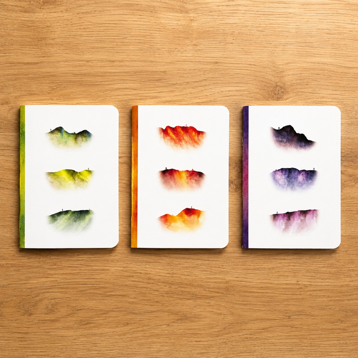 Three small notebooks with white covers are arranged side by side on a light brown wooden surface. Each notebook has three abstract watercolour mountain designs on the cover, with green, orange, and purple tones.