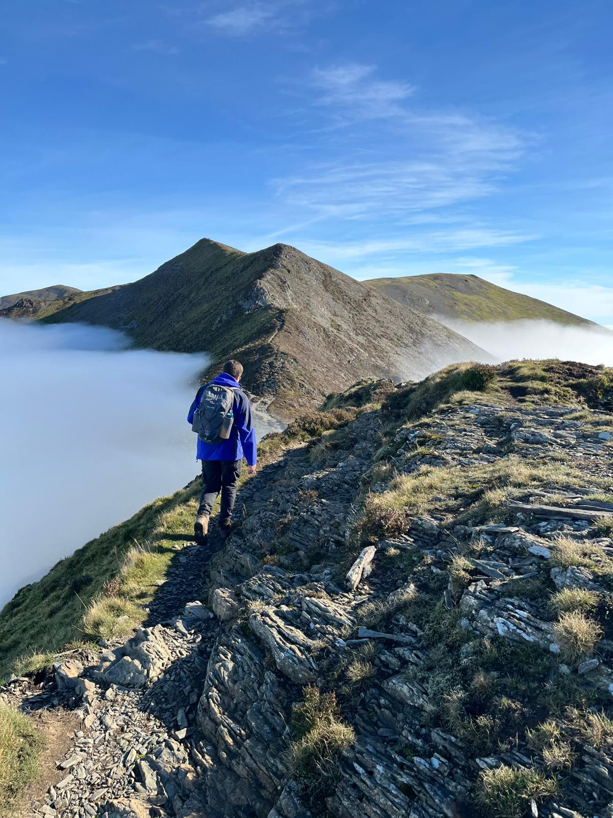 A hiker wearing a blue jacket and a gray backpack walks along a rocky ridge path between Whiteside and Hopegill Head with a sea of clouds below under a bright blue sky.